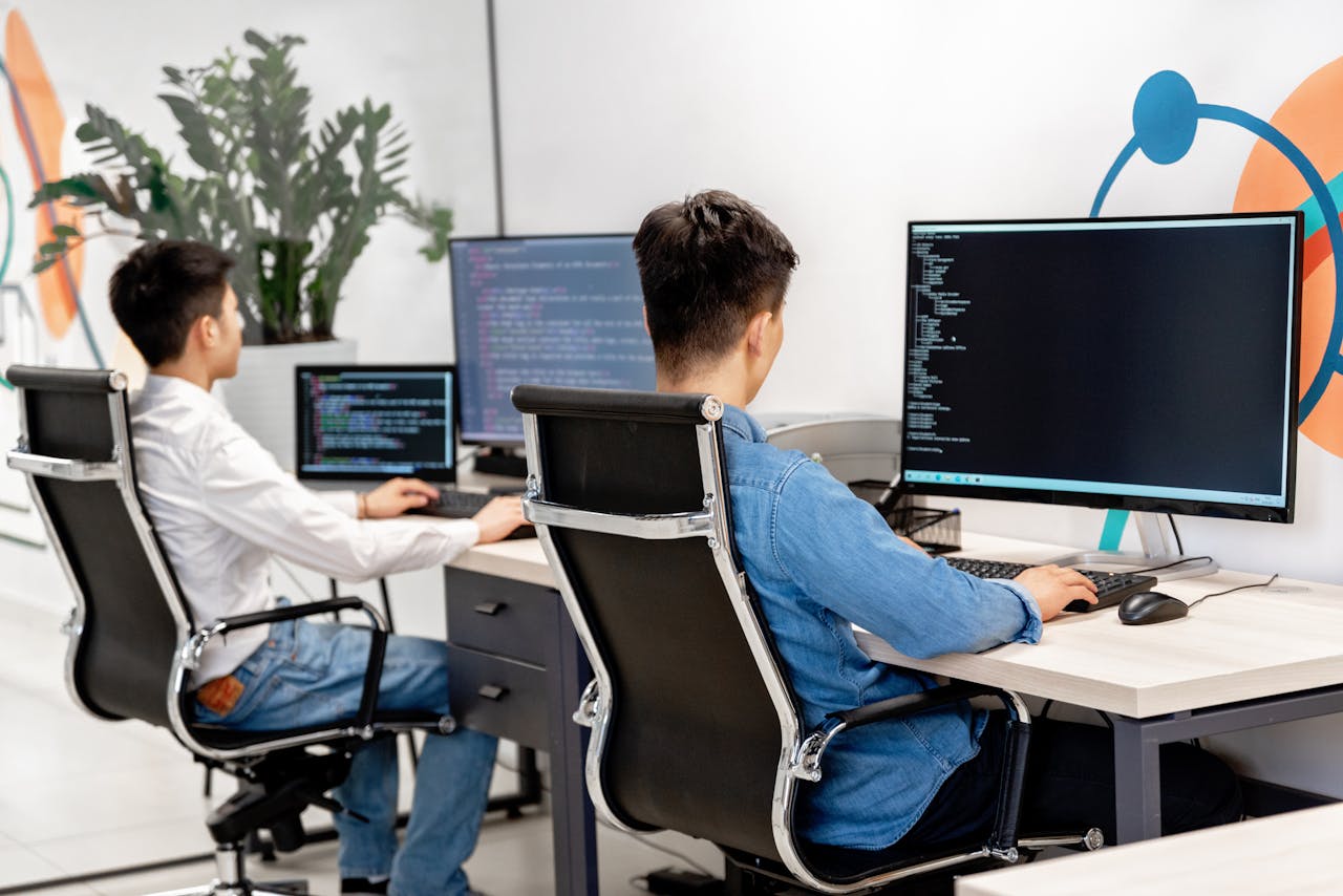 hero-img-01 Two male developers at desks programming in a modern office workspace with large monitors.